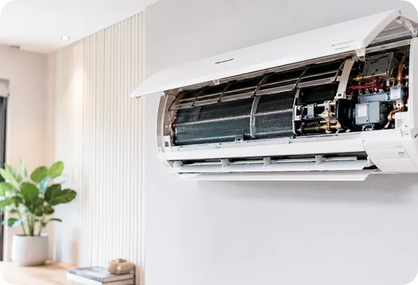 Wall-mounted air conditioner with the front cover open, exposing internal components. The room is modern with a plant on a wooden shelf in the background.