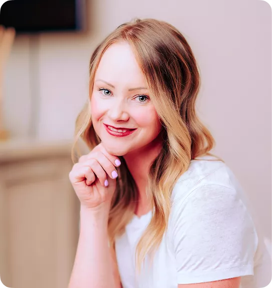 Smiling woman with long wavy blonde hair in a white top, resting her chin on her hand.