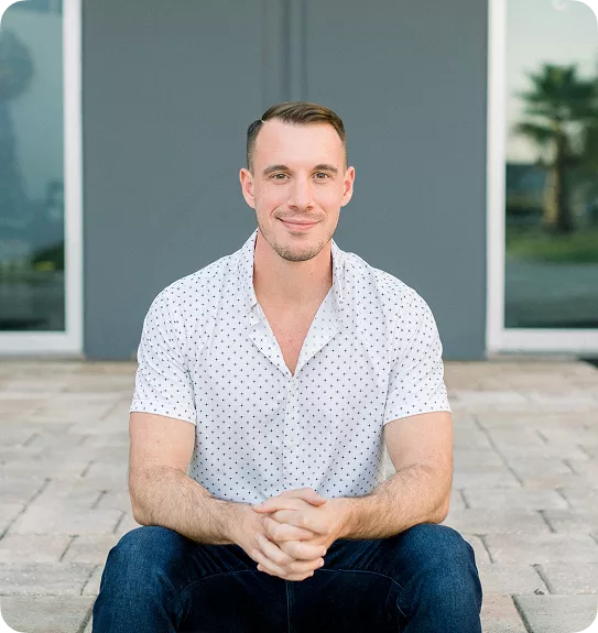 Smiling man with short brown hair in a white polka-dot shirt, sitting outdoors with hands clasped.